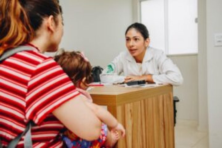 A imagem em destaque mostra uma médica, atrás da mesa, atendendo uma mulher com